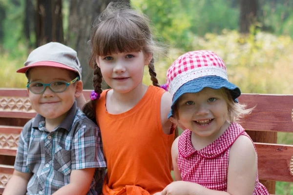 Szczęśliwy, że mały chłopiec i dwie dziewczyny siedzą na ławce i uśmiecha się słoneczny — Zdjęcie stockowe