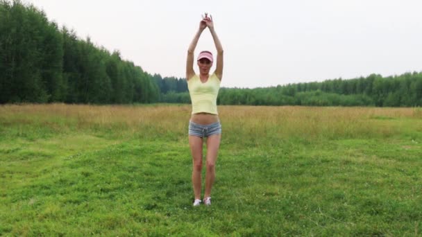 Красивая девушка в шортах и кепке танцует на зеленом лугу летним вечером — стоковое видео