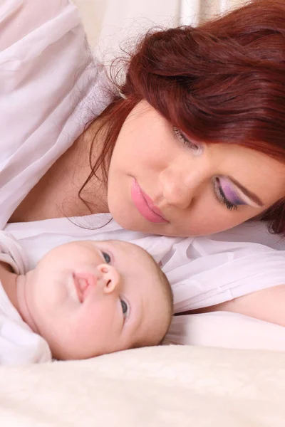 Junge Mutter im seidenweißen Kleid mit Baby auf dem Bett liegend — Stockfoto