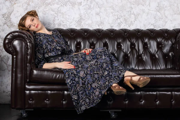 Young woman with hairdo in dress lies on black leather sofa — Stock Photo, Image