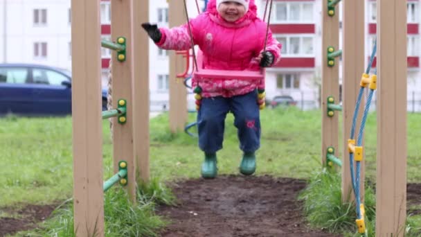 Маленькая девочка в теплой одежде качается на детской площадке и улыбается — стоковое видео