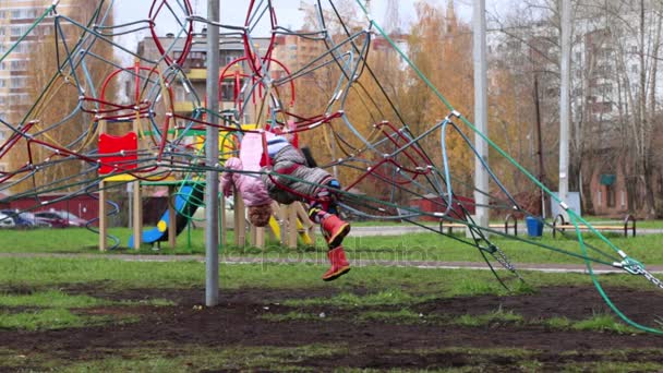 Маленька дівчинка і хлопчик в теплому одязі піднімаються і зависають на дитячому майданчику — стокове відео