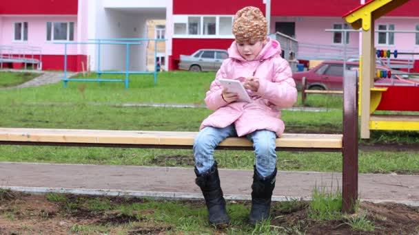 Маленькая девочка в теплой одежде сидит на скамейке и играет с планшетом на детской площадке — стоковое видео
