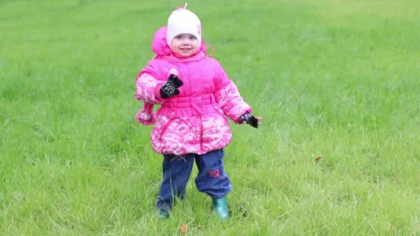 Klein meisje in warme kleding staat op gras en golven van de hand — Stockvideo