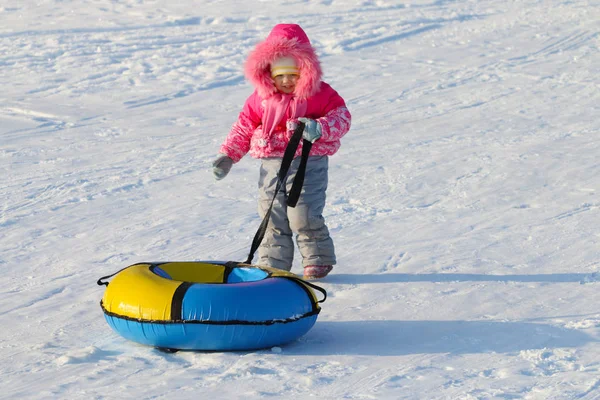 Niña feliz arrastra el tubo de nieve brillante en la nieve en el día de invierno —  Fotos de Stock