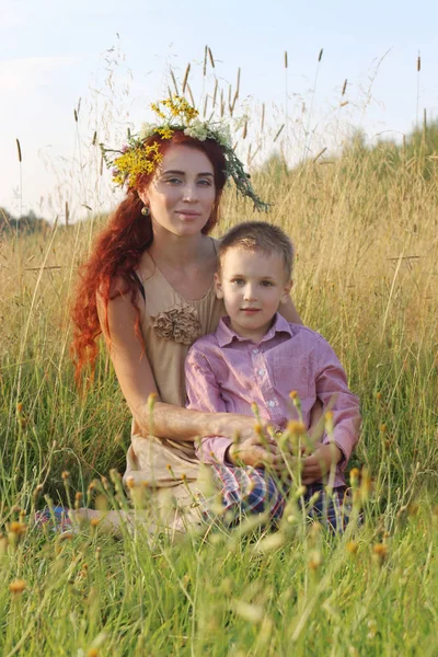 Happy woman in wreath and little son sit in dry grass at summer — Stock Photo, Image
