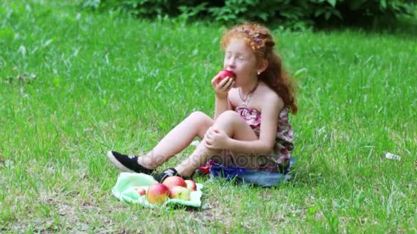 Little girl with red hair eats apple on lawn in green summer park — Stock Video