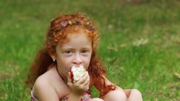 Niña come manzana en el césped en el verde parque de verano, de cerca — Vídeo de stock