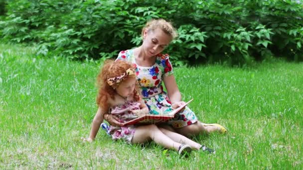 Klein meisje en haar moeder lezen boek op grasveld in groene zomer park — Stockvideo