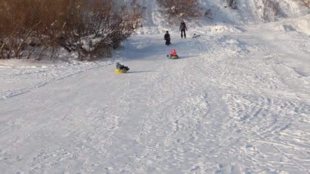 Маленькая девочка скользит на снегоходе с холма, двое неузнаваемых взрослых и ребенок на холме — стоковое видео