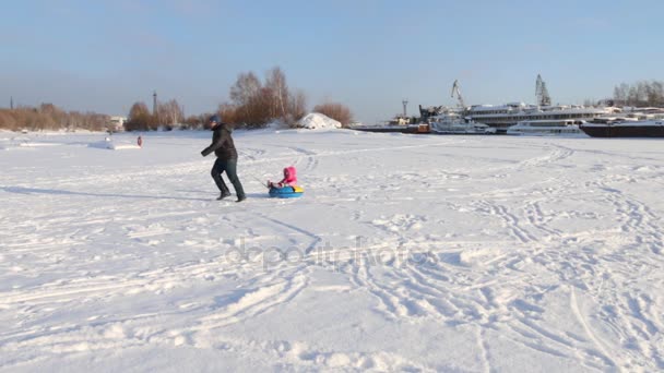 Happy father rolls his little daughter on snowtube at frozen river near ships — Stock Video