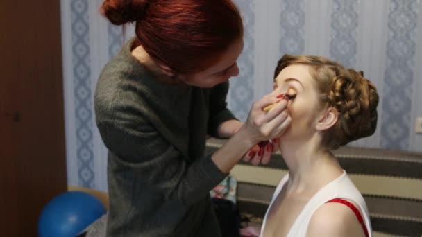 Макияж художник делает макияж для красивой женщины с прической в комнате — стоковое видео