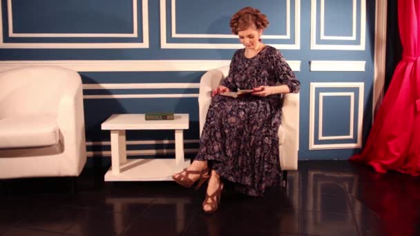 Mulher de vestido senta-se perto da mesa no quarto azul e lê livro — Vídeo de Stock