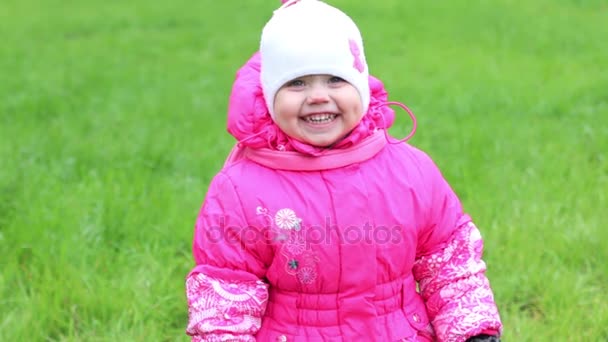 Маленькая девочка в теплой одежде стоит на траве, улыбается и смеется — стоковое видео