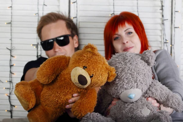 Chica de pelo rojo y hombre en gafas de sol se sienta con osos de juguete en studi —  Fotos de Stock