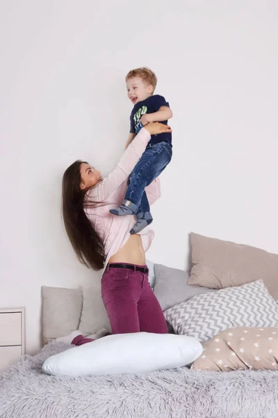 Joven madre juega con su pequeño hijo lindo en la cama con almohadas —  Fotos de Stock