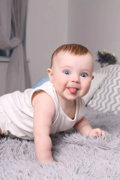 Little funny baby in white puts out tongue on bed with pillows i — Stock Photo, Image