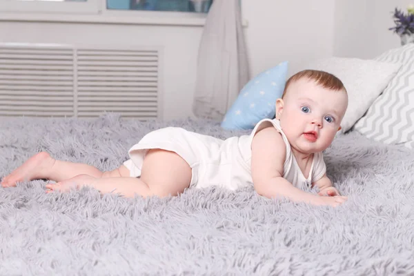 Little funny baby in white lies on bed with pillows in bedroom — Stock Photo, Image