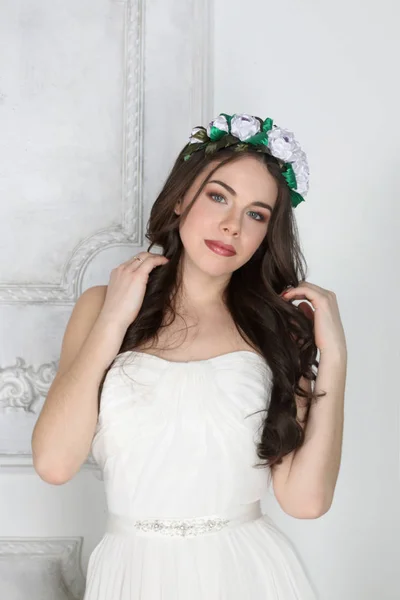 Young cute brunette in wreath and in white dress poses in studio — Stock Photo, Image