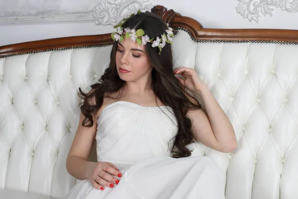 Young cute brunette in wreath and in white dress poses on couch — Stock Photo, Image