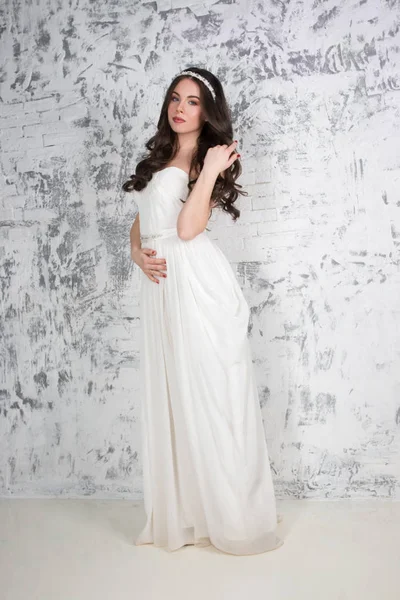 Pretty young brunette in long white dress poses near wall in whi — Stock Photo, Image