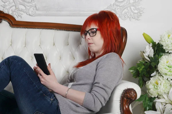 Ładna dziewczyna w okularach siedzi na białej kanapie z smartphone w whi — Zdjęcie stockowe