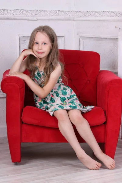 Happy barefoot little girl in dress sits in armchair in white ro — Stock Photo, Image