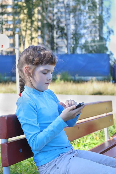 Девушка в синем сидит со смартфоном на скамейке на детской площадке — стоковое фото