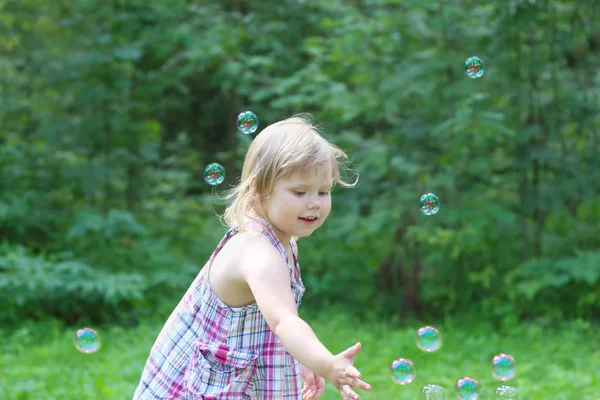 Happy blonde little girl catches soap bubbles in summer green pa — Stock Photo, Image