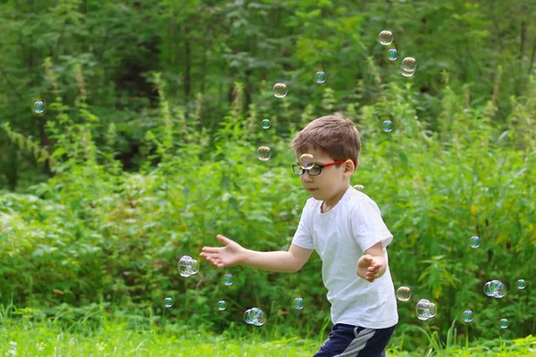 Happy little boy in glasses catches soap bubbles in summer green — Stock Photo, Image