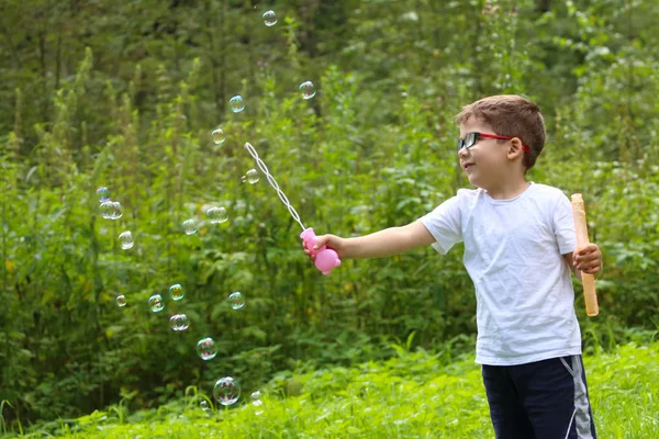 Glimlachend jongetje in glazen speelt met zeepbellen in groene f — Stockfoto