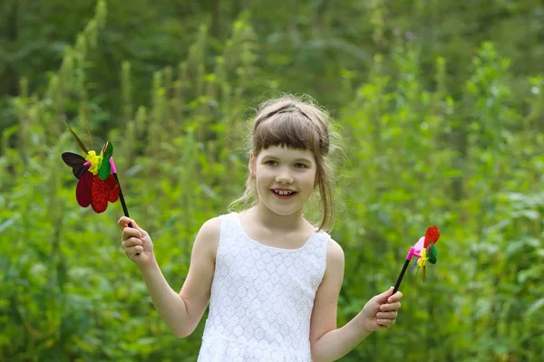 Kleines Mädchen in Weiß hält zwei Windmühlen und lächelt im Sommer. — Stockfoto