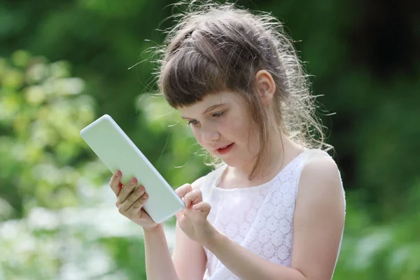 Little smiling girl in white dress plays with tablet pc in green — Stock Photo, Image