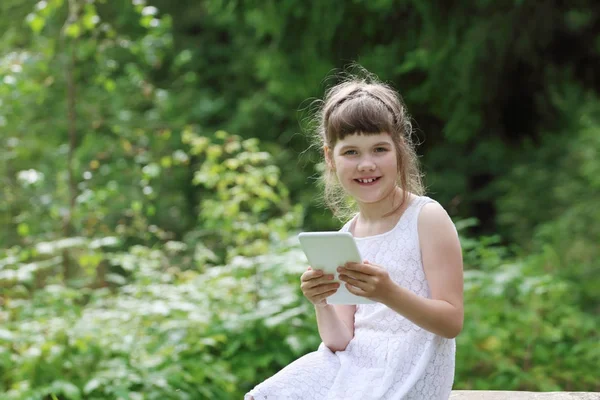 Девочка в белом платье улыбается и держит в руках планшетный компьютер в зеленом — стоковое фото