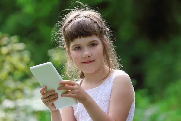 Маленькая улыбающаяся девочка в белом платье держит белый планшет ПК в парке — стоковое фото