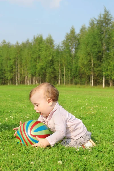 Cute happy baby with ball sits on green grass on summer field — Stock Photo, Image