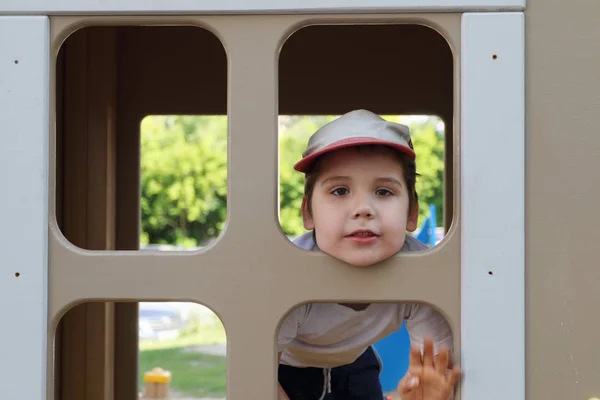 Little boy in cap looks into window in wooden house — Stock Photo, Image