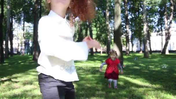Дві красиві дівчата ловлять мильні бульбашки в зеленому літньому парку — стокове відео
