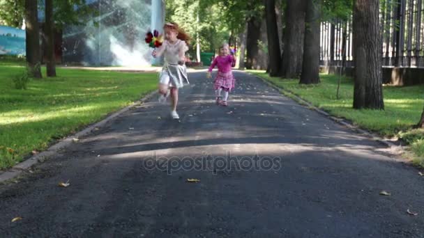 Duas meninas felizes correm com moinhos de vento no parque de verão verde — Vídeo de Stock