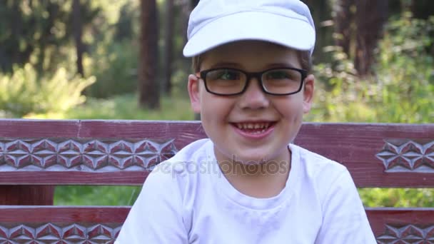 Niño feliz en gafas y gorra se ríe en el banco en el parque de verano — Vídeo de stock