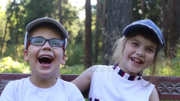 Щасливий хлопчик і дівчинка в шапках сміються на лавці в літньому парку — стокове відео