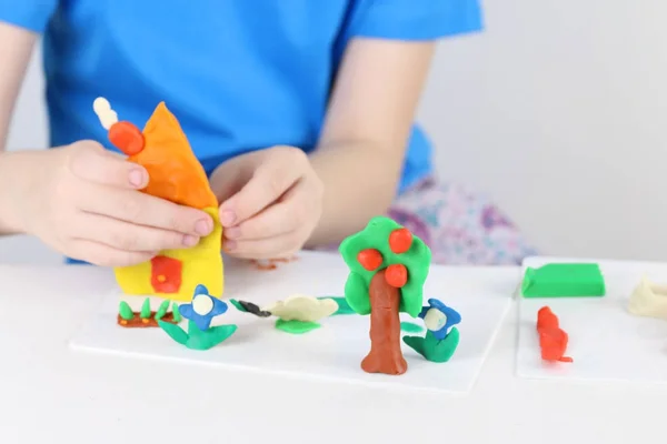 Child hands molding house, tree, flowers from plasticine on tabl — Stock Photo, Image