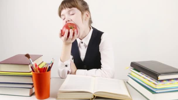Schoolgirl sits with book and eats red apple at white table — Stock Video