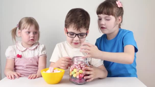 Трое счастливых детей достают зефир из банки и едят на столе в белой студии. — стоковое видео