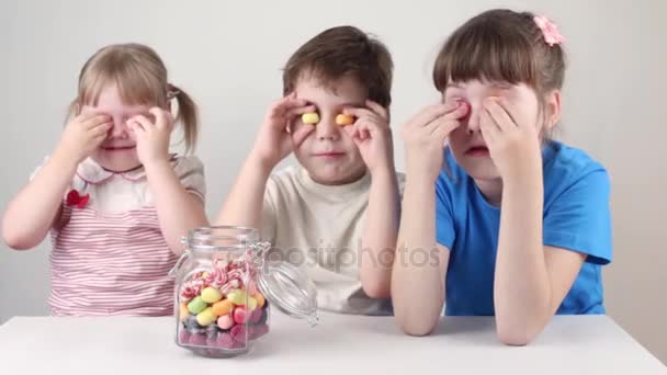 Tres niños felices juegan con caramelos cerca del frasco en la mesa en el estudio blanco — Vídeos de Stock