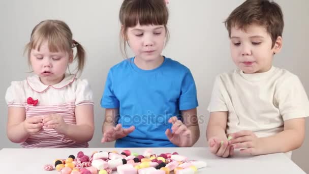 Tres niños felices comen caramelos y se sientan a la mesa en un estudio blanco — Vídeos de Stock