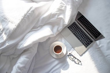 Laptopun üst görüntüsü ve sabah yumuşak güneş ışığıyla yatakta sıcak kahve..