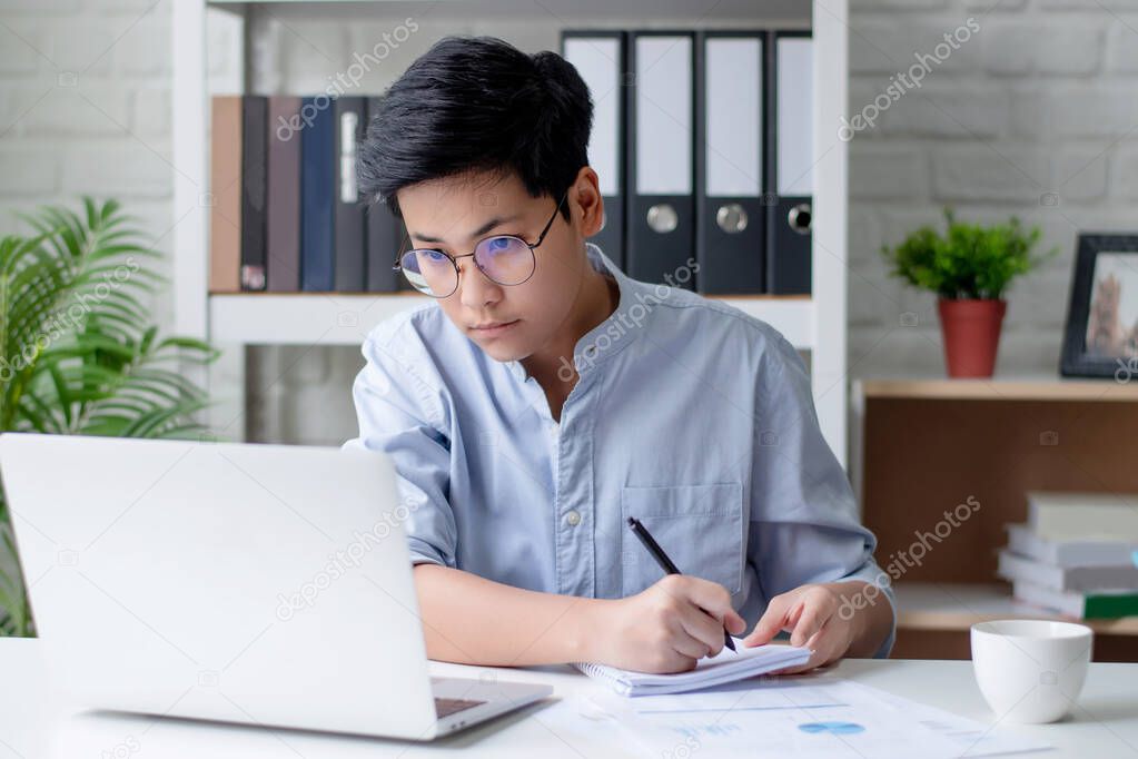 Young Asian businessman sitting at a desk is writing notes in a notebook using a laptop computer at home, working at home.