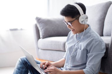 Genç Asyalı insanlar evlerindeki oturma odasında, koltuğun yanında dizüstü bilgisayarda oturup müzik dinlerken oturup çalışıyorlar..