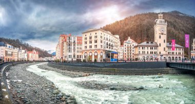 Evlerin, otellerin, dükkanların, Mzymtu Nehri 'nin, belediye kulesinin ve yağmurlu bir günde Rosa Khutor, Sochi' deki dağların karlı tepelerinin manzarası..